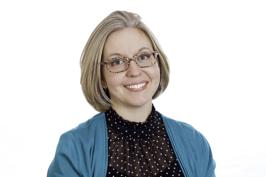 This Brian Charles Steel photo is a headshot of a white woman with blonde hair.  She is wearing glasses, a light blue green sweater and a black blouse with white polka dots.  She is centered in the frame, and the background is completely white.  The woman is lit with a Rembrandt style of lighting. 